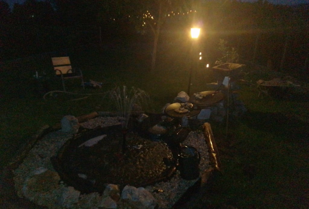 A dark nighttime scene showing a garden fountain illuminated by a warm lamp beside it,
