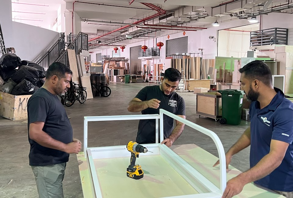 Professional carpenters assembling a white custom furniture frame using a power drill in a workshop.