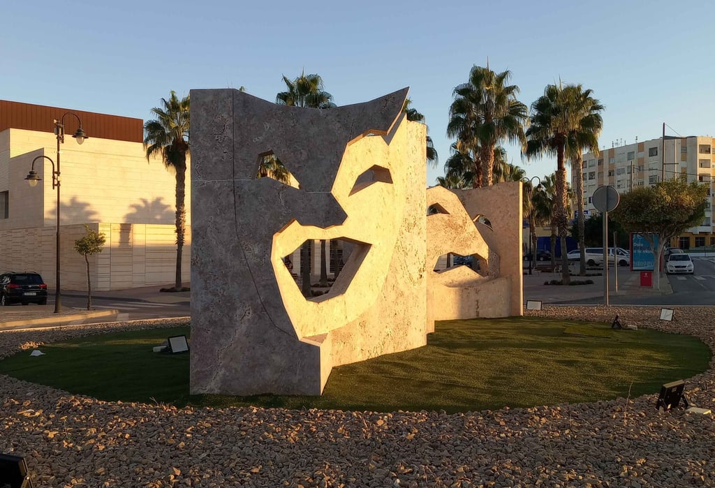 Large stone theater mask sculpture on a public roundabout with palm trees and city buildings.