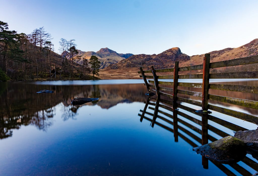Rustic wooden fence reflecting in the calm blue water of a lake with scenic mountain peaks.