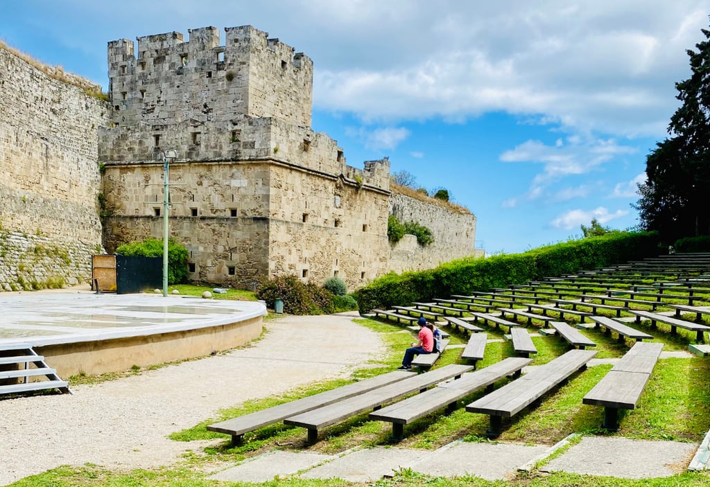 Outdoor stone amphitheater and historic castle walls in Rhodes, Greece, under a blue sky.