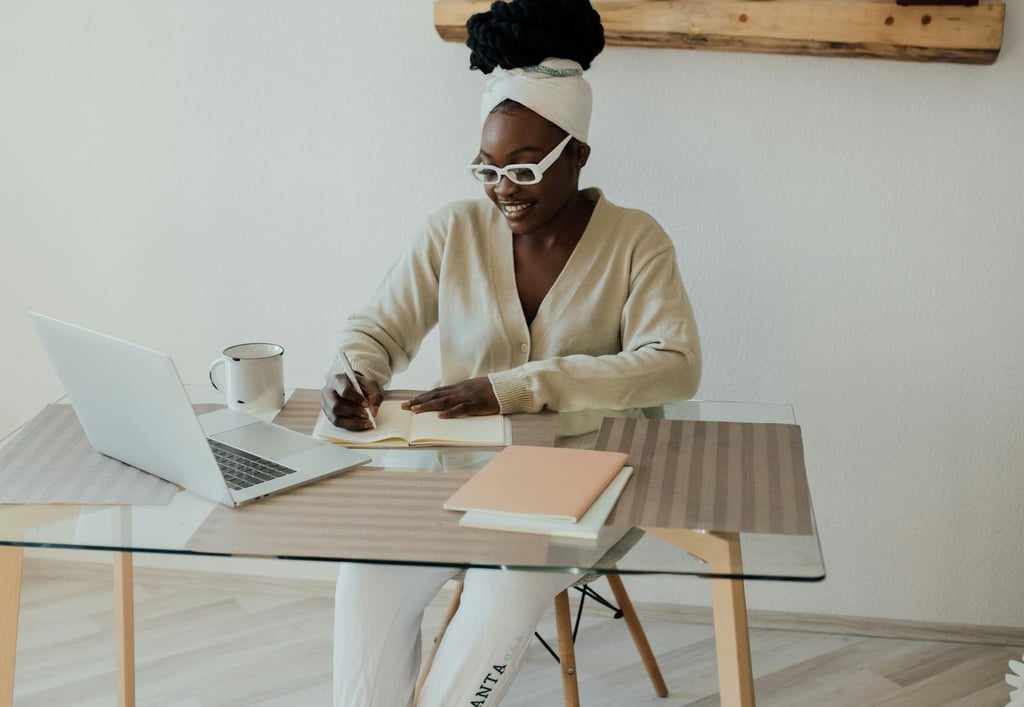 a woman sitting at a desk with an open laptop computer and writing in a notebook