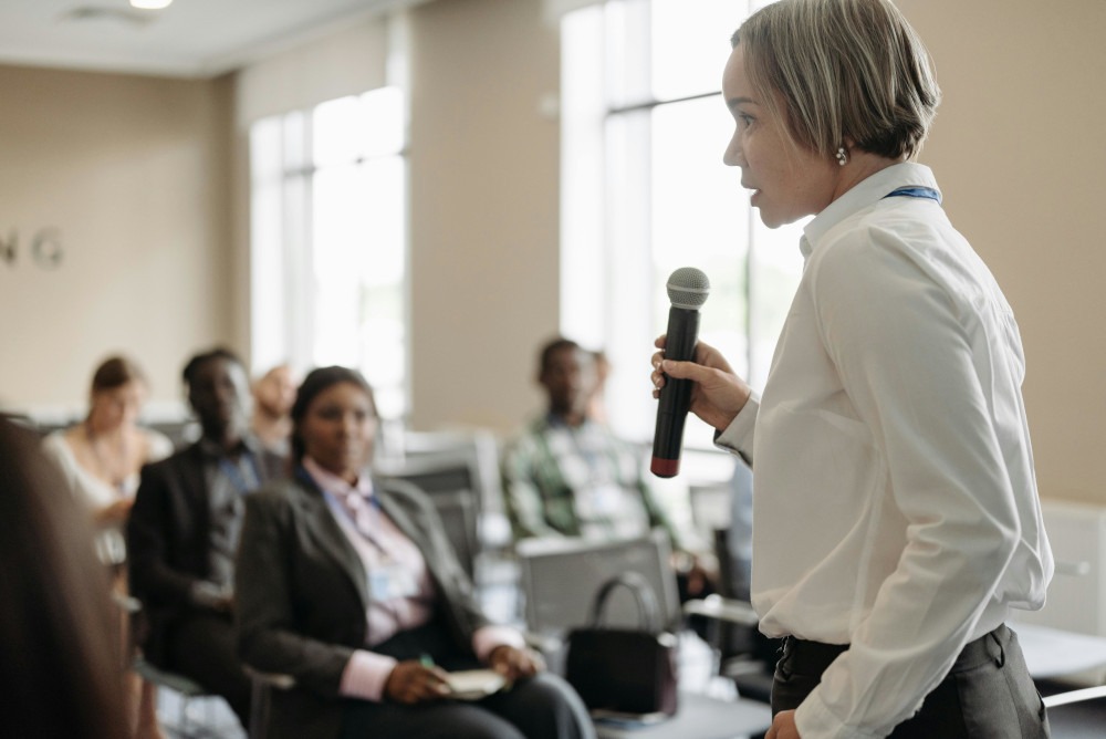 Professional female speaker holding a microphone and addressing a diverse audience at a business conference.