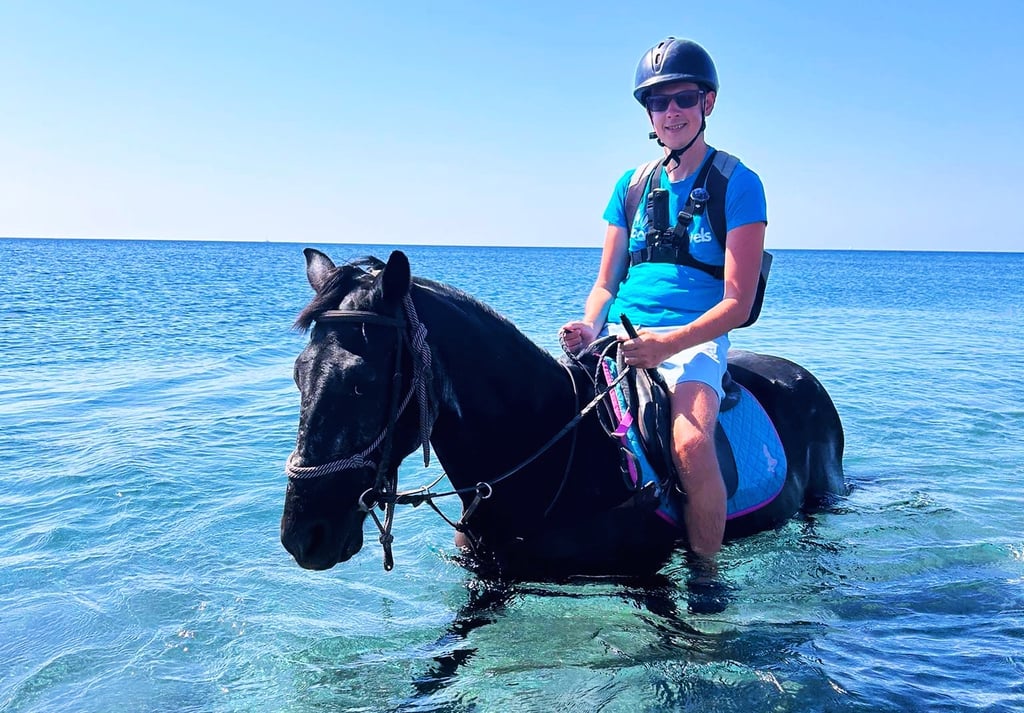 looneytravels riding a black horse through clear blue ocean water during a summer beach horse trek.