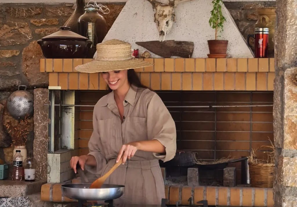 Smiling woman in a straw hat cooking outdoors on a gas stove in a rustic stone kitchen.