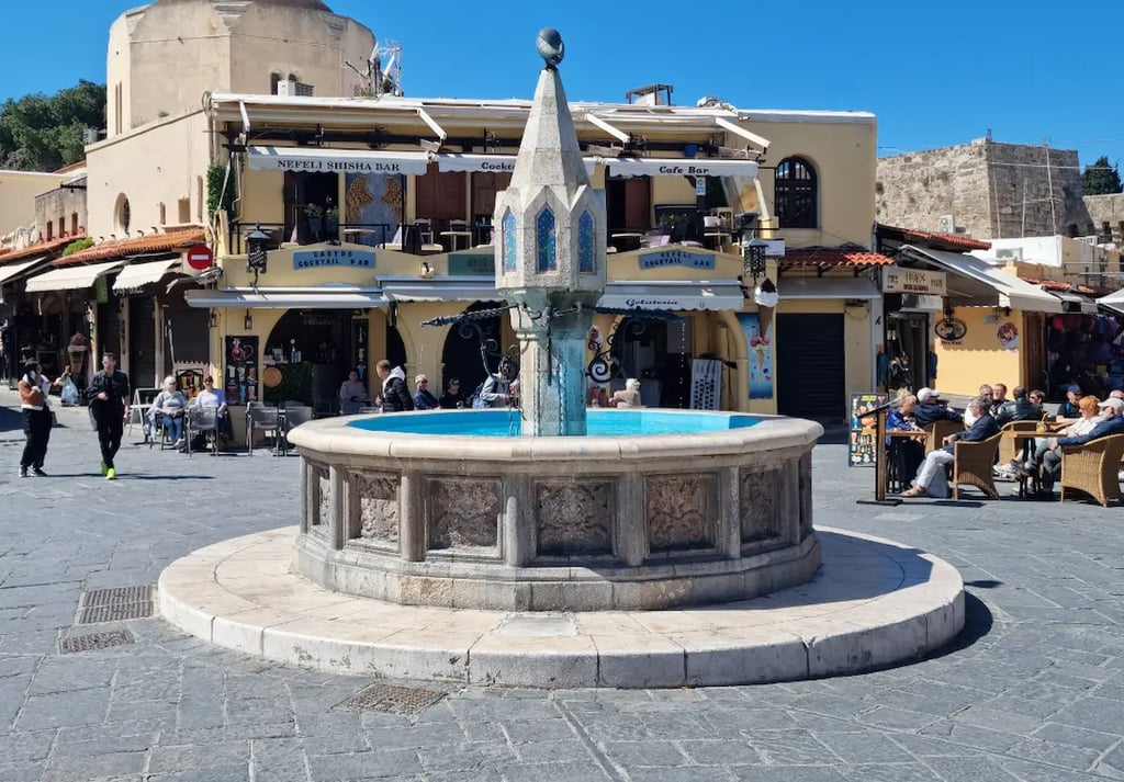 Historic Castellania Fountain in Rhodes Old Town square with cafes and a domed mosque background.