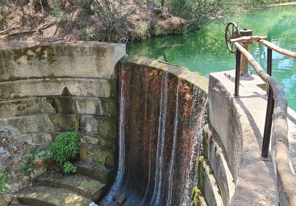 A small concrete dam and waterfall flowing into a turquoise pond surrounded by lush green trees.