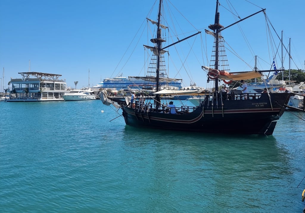 A vintage-style wooden pirate ship tour boat sailing in the blue waters of a Mediterranean harbor.
