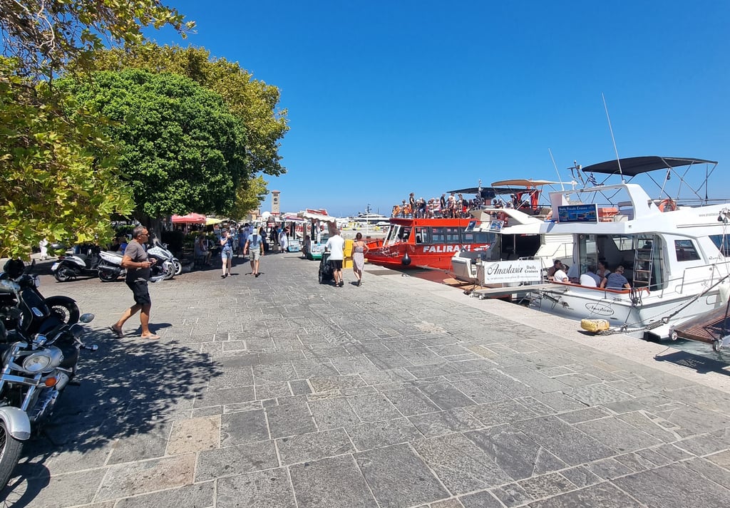 Crowded Mandraki Harbor in Rhodes Greece with tourist boats moored along the stone promenade under a blue sky.