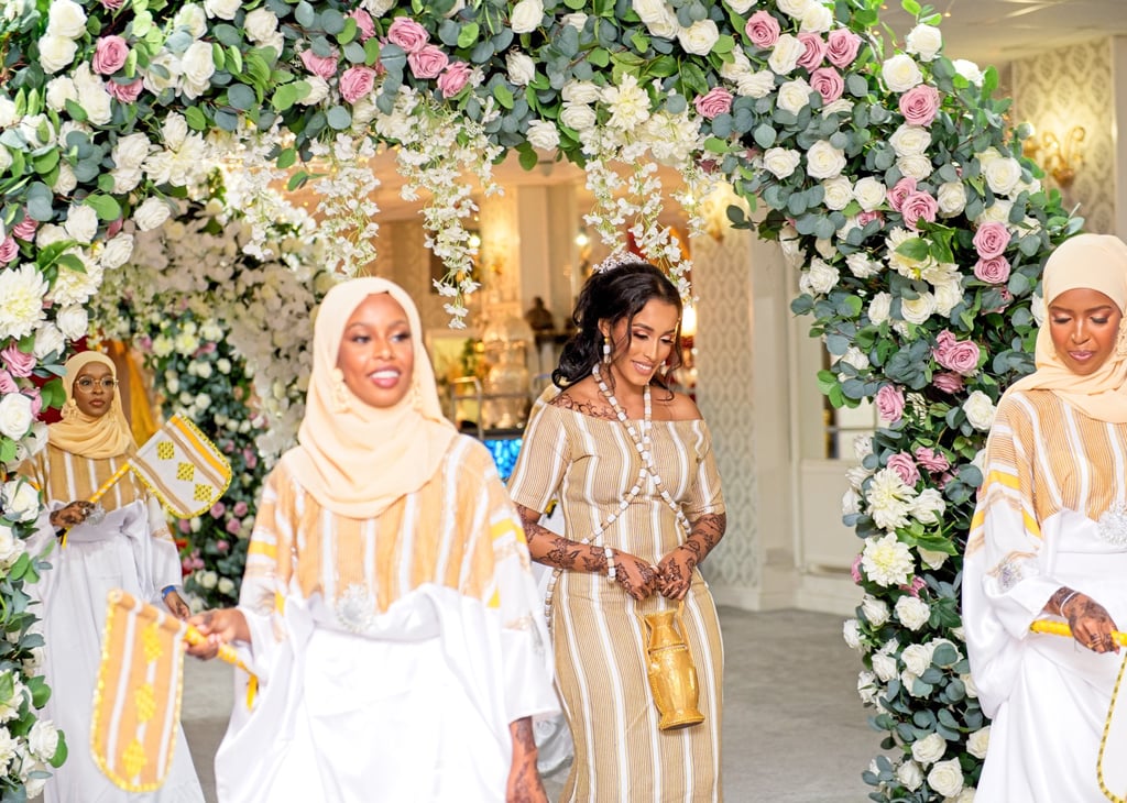 Somalian bride & bridesmaids all dressed in traditional white & cream outfits dancing and laughing , floral arch framing them