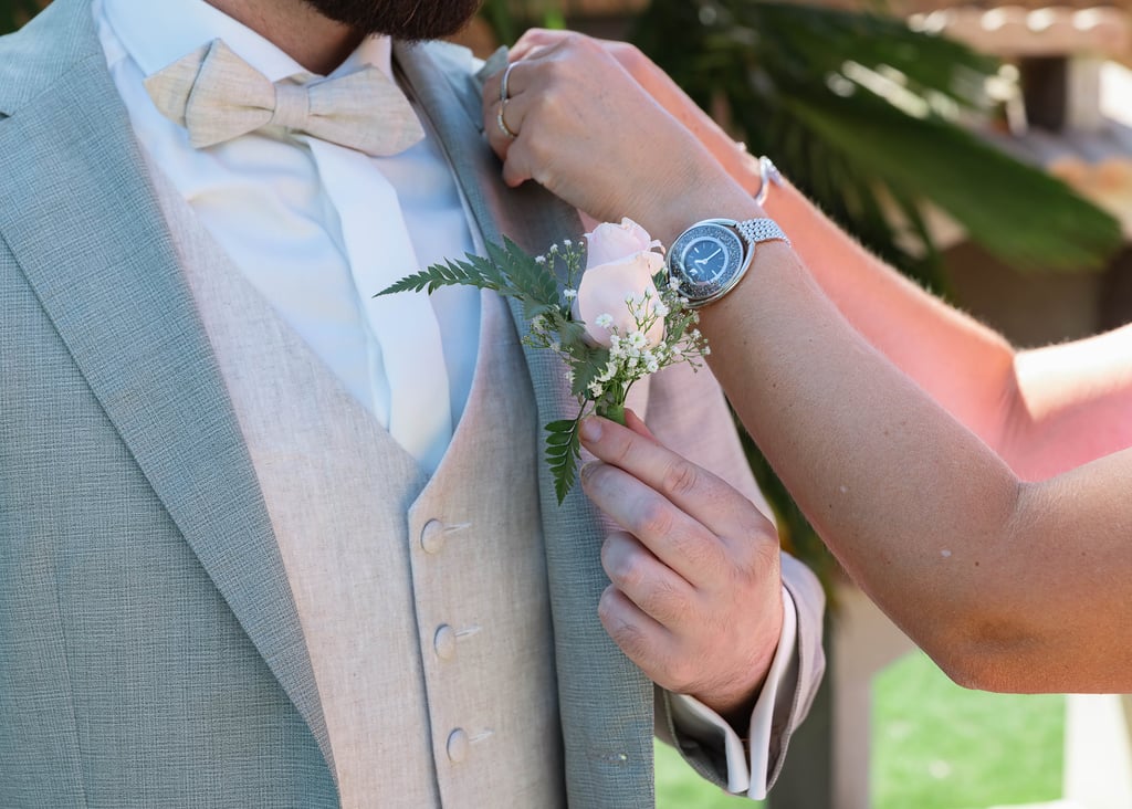 Une femme attache une boutonnière de rose rose au costume de mariage en lin gris clair du marié.