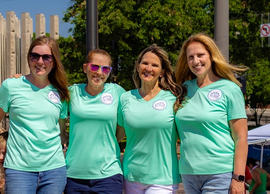 Four women smiling outdoors in matching mint green yoga t-shirts during a community wellness event.