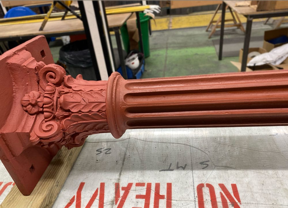 A red primed Victorian metal column and capital lying on table in industrial setting.