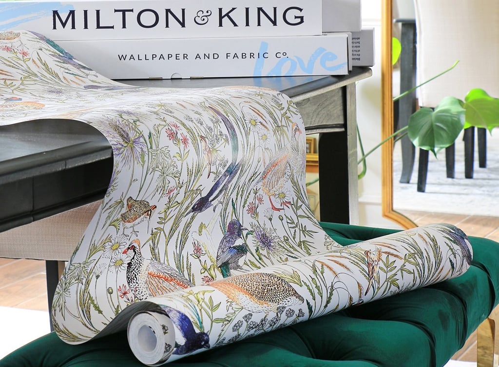 Milton and King floral bird wallpaper roll draped over a green velvet ottoman in a bright room.