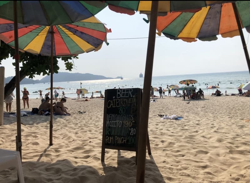 Sonniger Patong Beach in Phuket mit bunten Sonnenschirmen und einer Getränkekarte im Sand