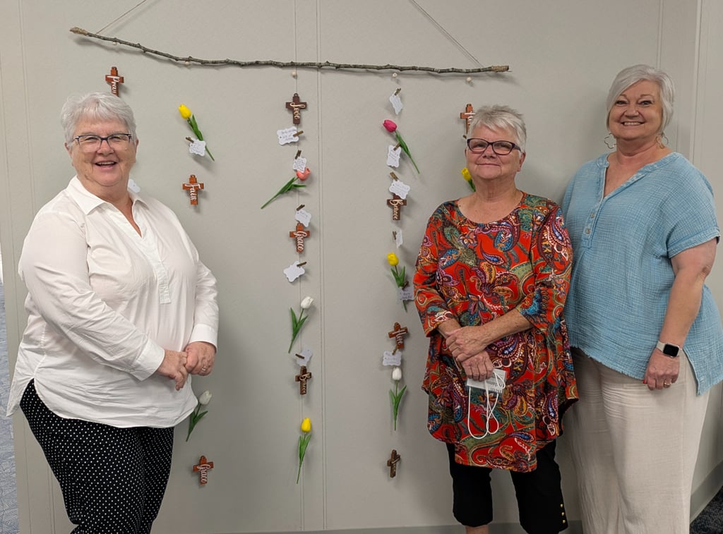 Three smiling women stand beside a hanging prayer wall display with wooden crosses and tulips.