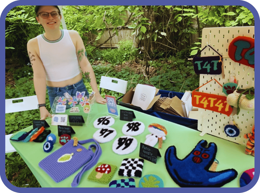 Artist selling handmade punch needle crafts and textile accessories at an outdoor market stall.