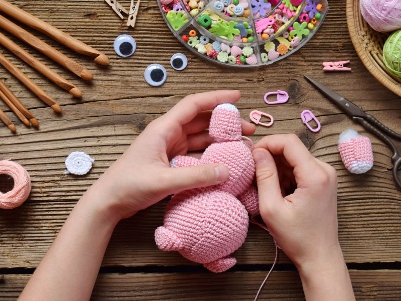 Manos ensamblando un amigurumi rosa tejido a crochet, rodeado de ganchos y estambre.
