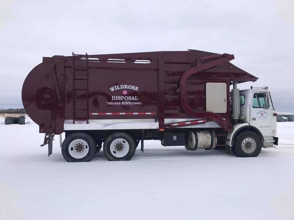 A maroon Wildrose Disposal front-load garbage truck parked in a snowy field for waste management services.