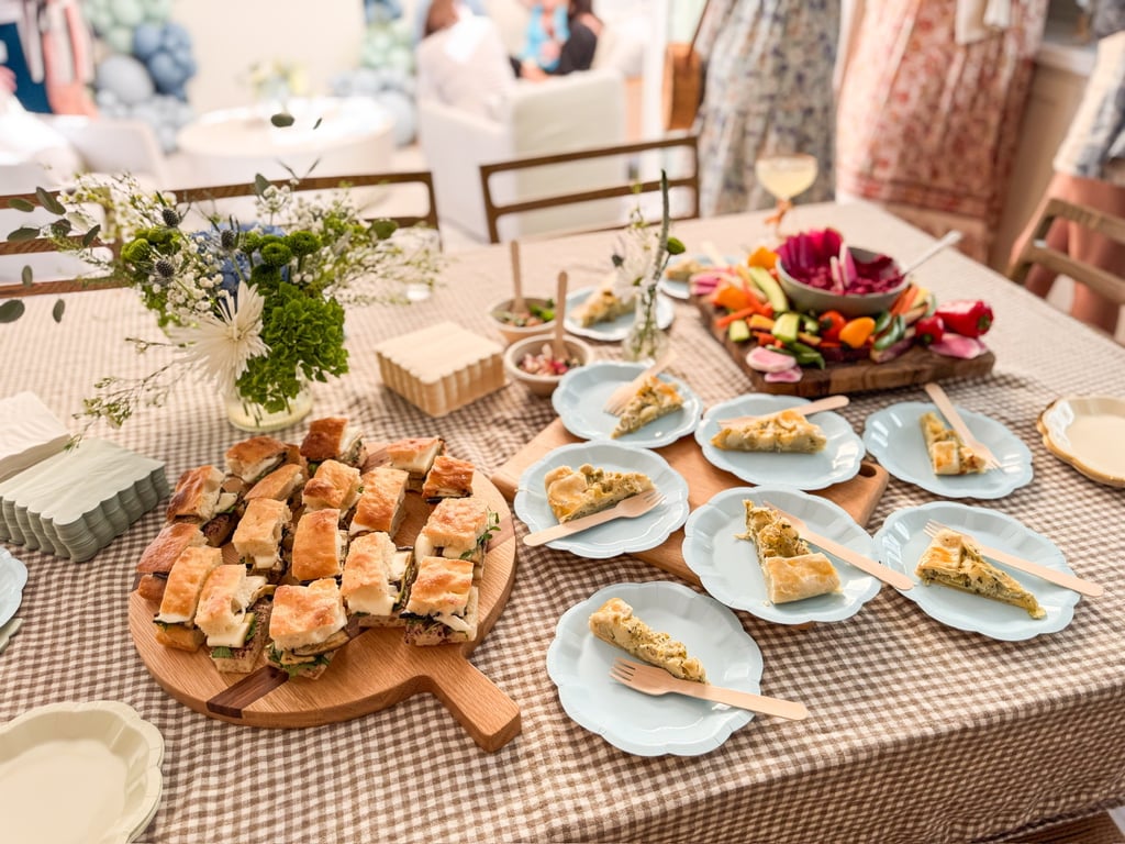 A catered party table with mini sandwiches, savory quiche slices, and fresh vegetable crudité.