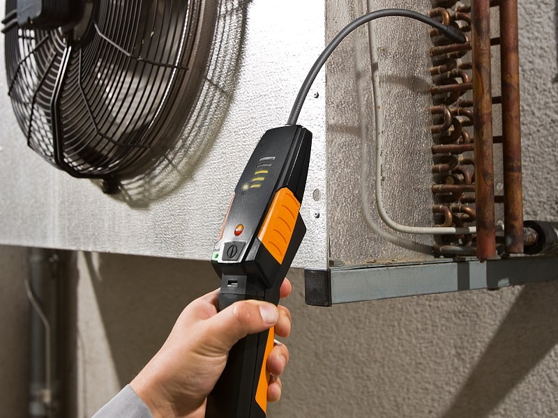 A hand holding a handheld electronic refrigerant leak detector near a freezer unit condenser coil.