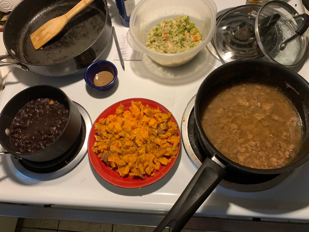 A variety of healthy home-cooked dishes including black beans, sweet potatoes, and vegetable rice on a white stove.
