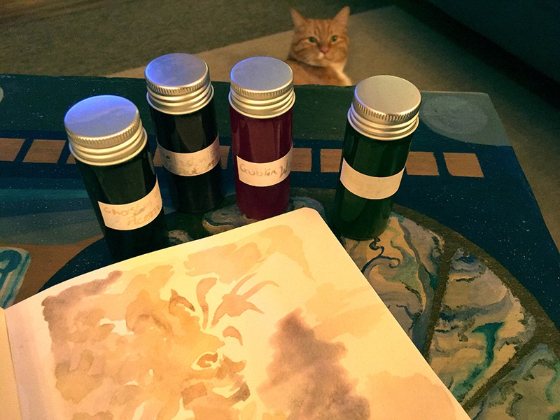 An open sketchbook with a fantasy watercolour illustration on a table surrounded by inks and a cat.