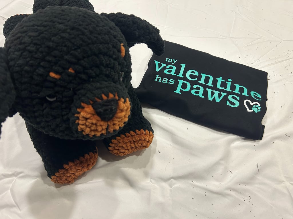 a folded tshirt that reads "my valentine has paws" sits beside a stuffed rottweiler