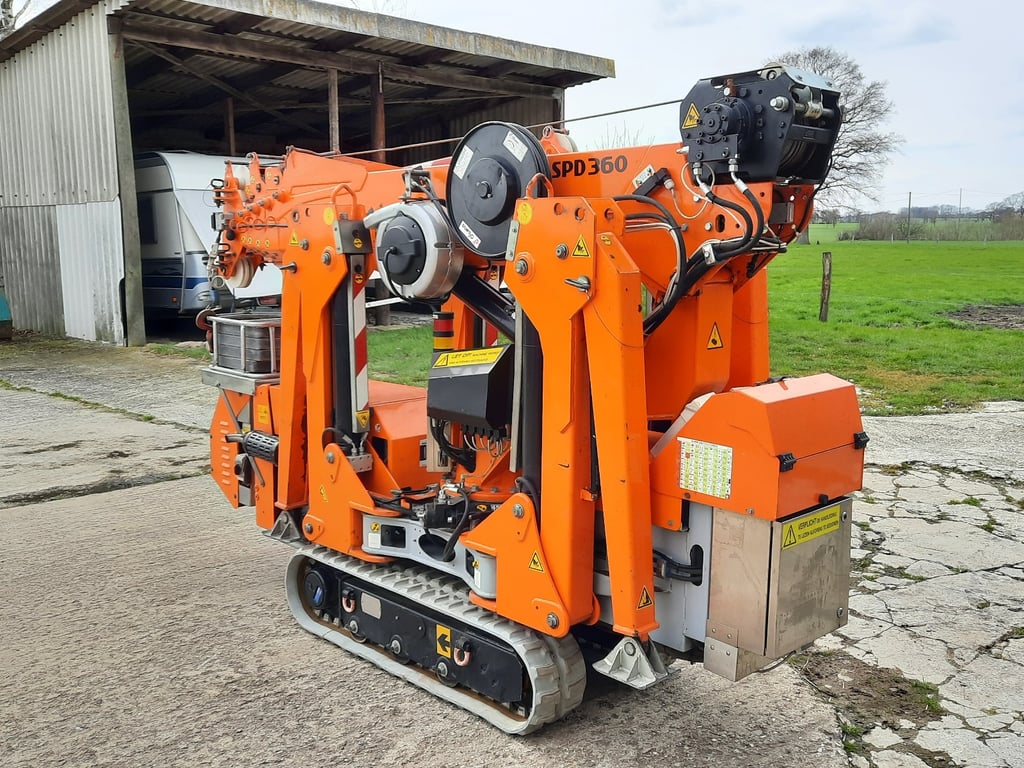 Kompakter oranger Minikran SPD360 auf Raupenketten, zusammengefaltet vor einer Scheune.