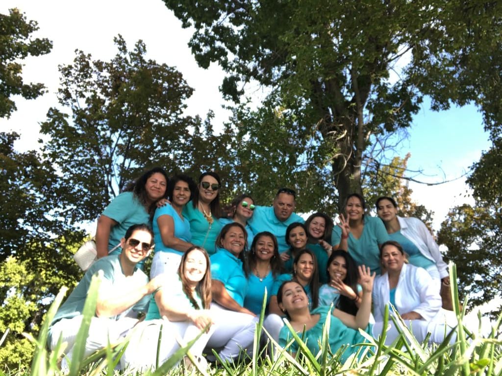 A smiling dental staff group photo outdoors wearing teal scrubs and white pants for a team building event.