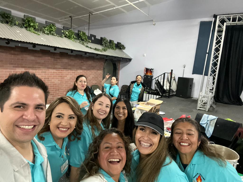 A cheerful group of event volunteers in matching blue shirts posing for a group selfie indoors.
