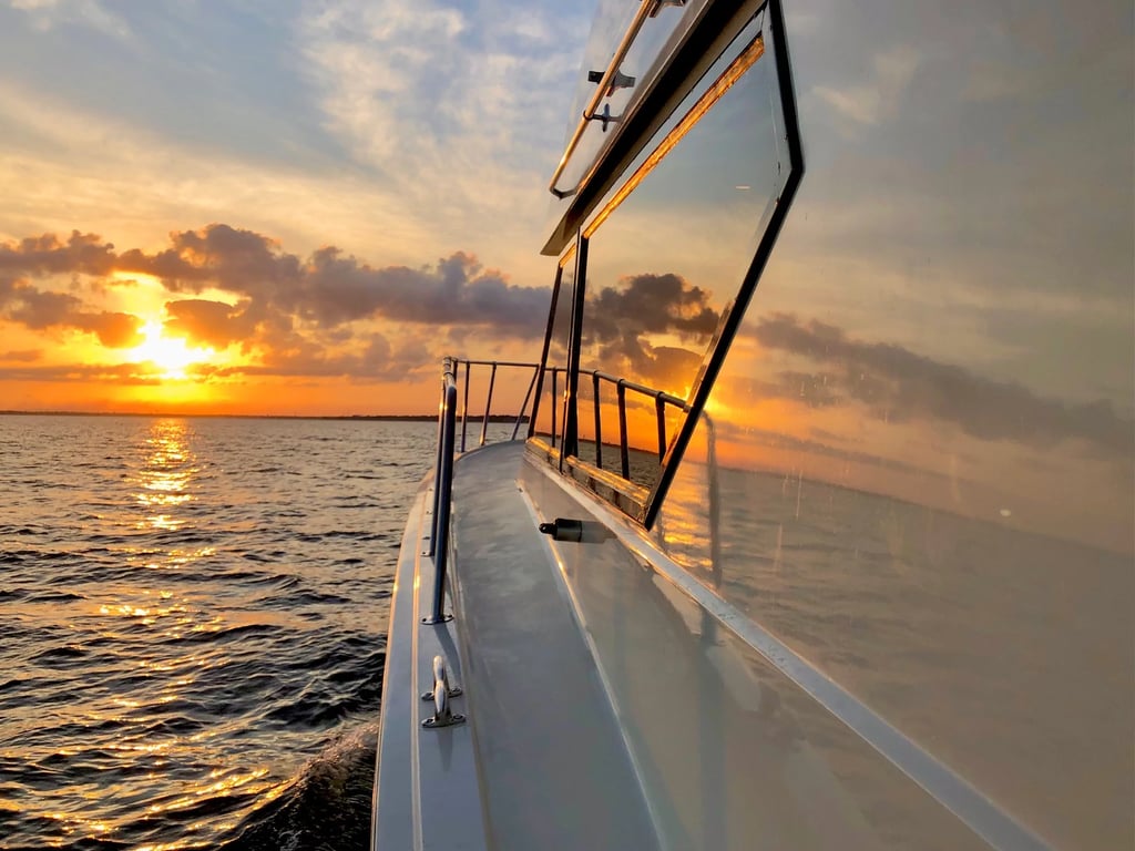 A luxury yacht cruising on the ocean at sunset with orange clouds reflecting on the water and cabin glass.