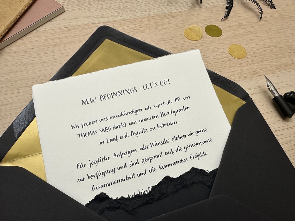 A Thomas Sabo announcement letter in a black and gold envelope on a wooden desk with a fountain pen.
