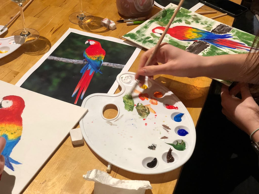 A person using a paint palette and brush to create a colorful scarlet macaw parrot acrylic painting.