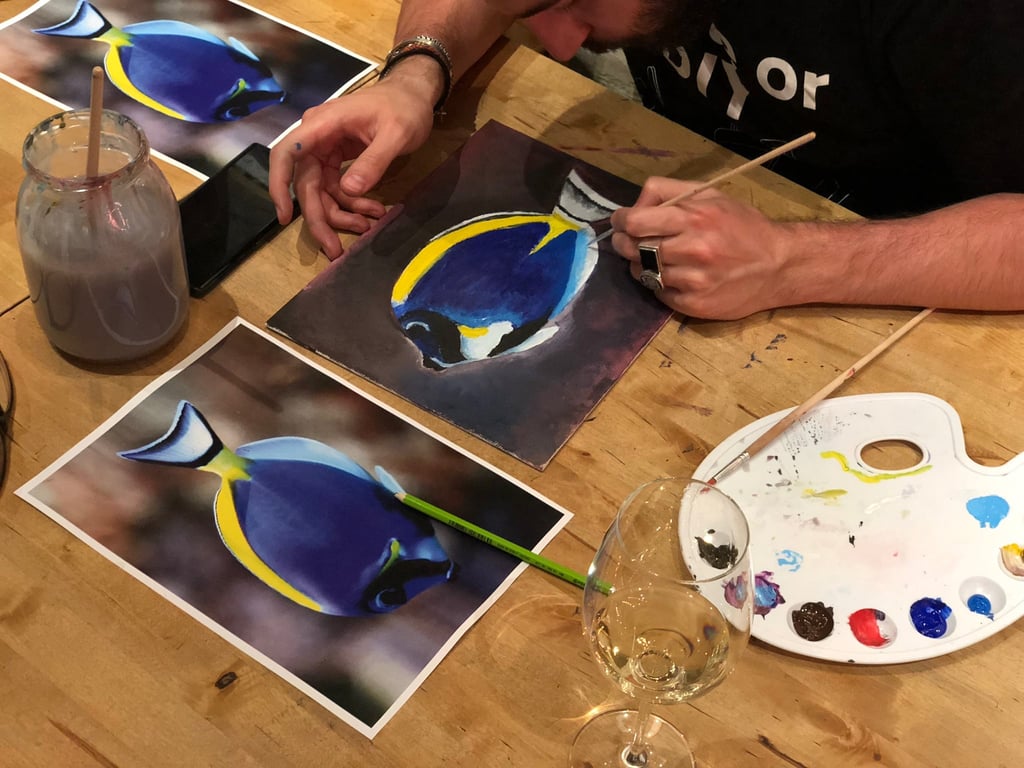 An artist using a paintbrush to create a realistic oil painting of a blue tang fish on a wooden table.