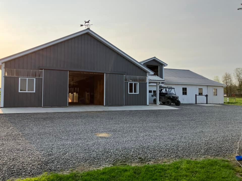 A modern charcoal grey metal horse barn with sliding doors and a white farm building on a gravel driveway.