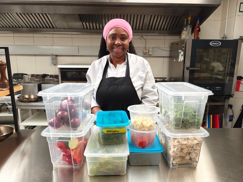 Chef Viviane smiling behind multiple labelled transparent prep containers filled with vegetables on a stainless steel.