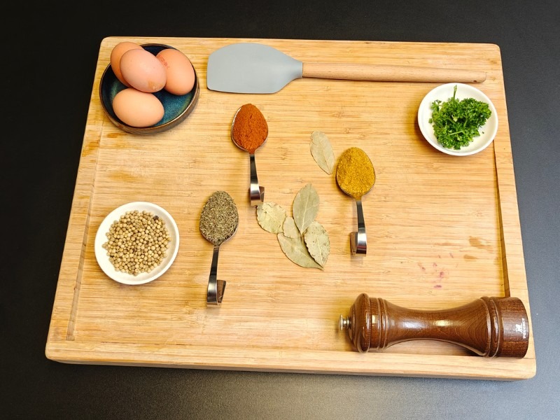 Overhead view of spices, eggs, fresh herbs and a pepper mill arranged on a bamboo chopping board on a dark