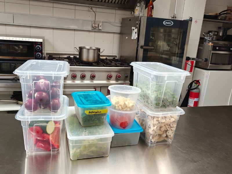 Multiple labelled transparent prep containers filled with vegetables and ingredients lined up on a stainless steel kitchen.