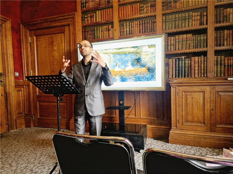 A man in a suit giving a presentation in a classic library with a digital screen showing artwork.