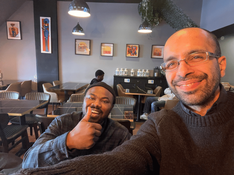Two smiling men taking a selfie inside a modern restaurant with artwork and pendant lighting.