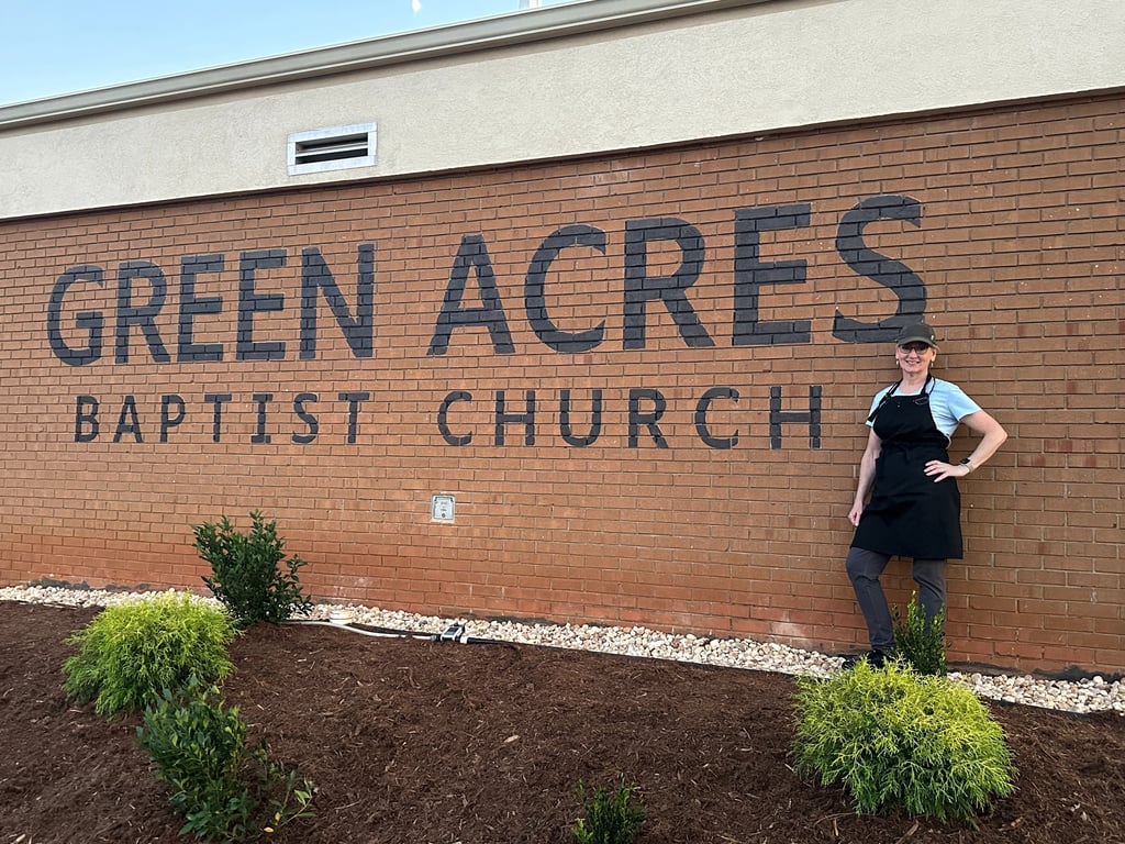 A smiling woman in an apron stands before the Green Acres Baptist Church brick sign.