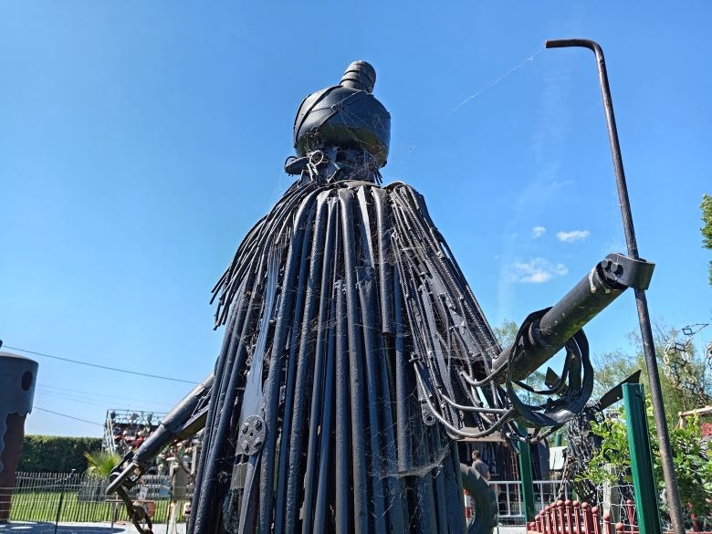 A tall black metal sculpture of a robed figure with a staff made from recycled pipes and scrap metal.