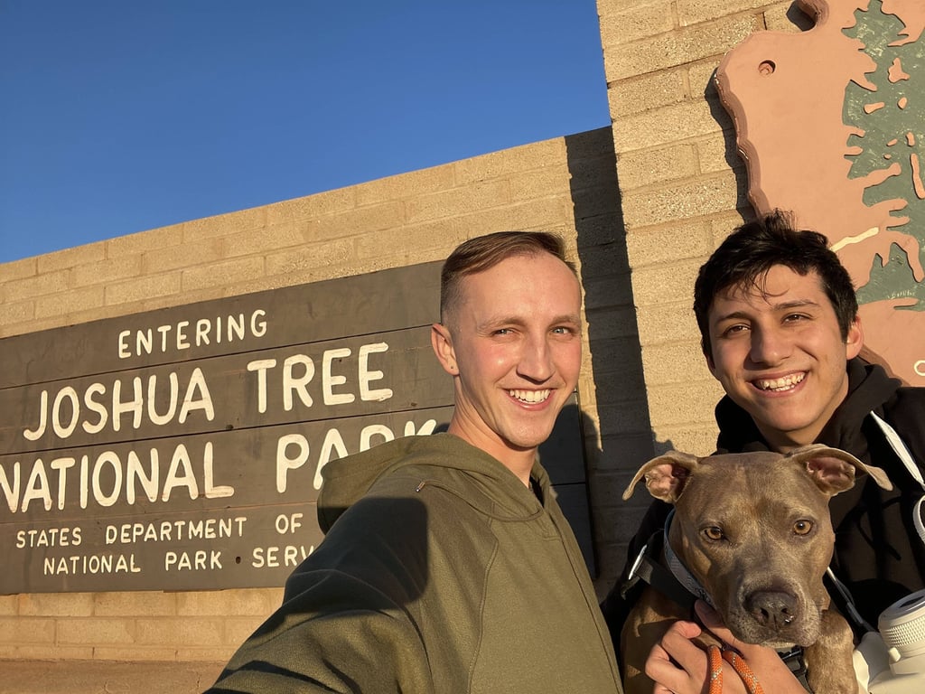 Two people and a dog smile in front of the Joshua Tree National Park entrance sign on a sunny day. 