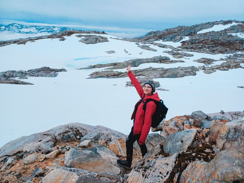 A female hiker in a red jacket standing on snowy mountain rocks during a winter trekking in Norway.