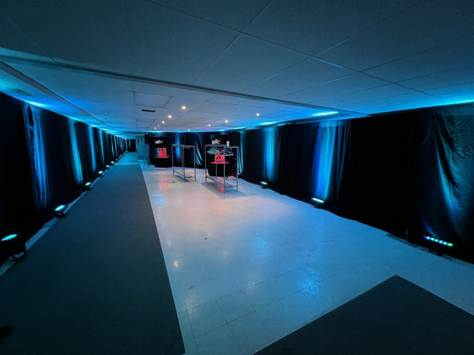 Professional event space with black drapes, blue LED uplighting, and clothing racks in a wide hallway.