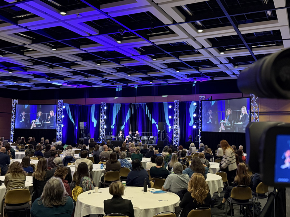 Audience at a professional conference seated at round tables watching a panel discussion on large screens.