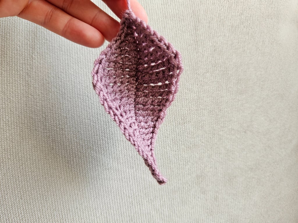 Hand holding a small purple crochet leaf pattern made from wool yarn against a neutral fabric background.