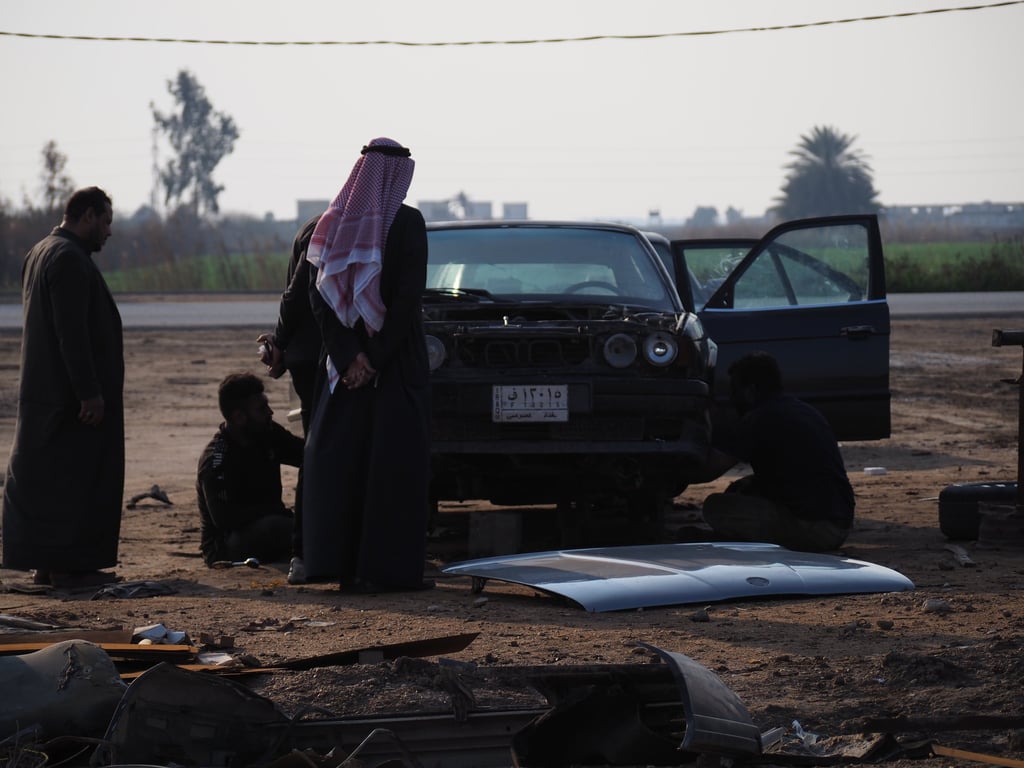 a group of Arab men in traditional clothes overlooking the dismantling of an old BMW