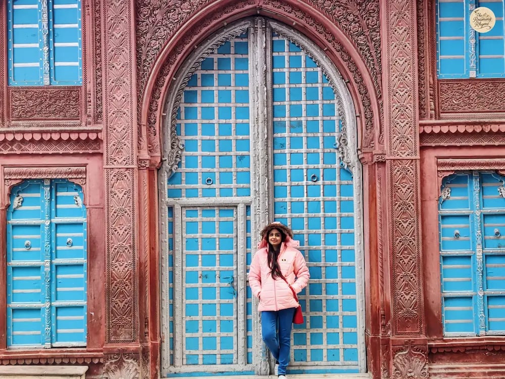 Person strolling past historic havelis in Bikaner with intricately carved doors and windows.